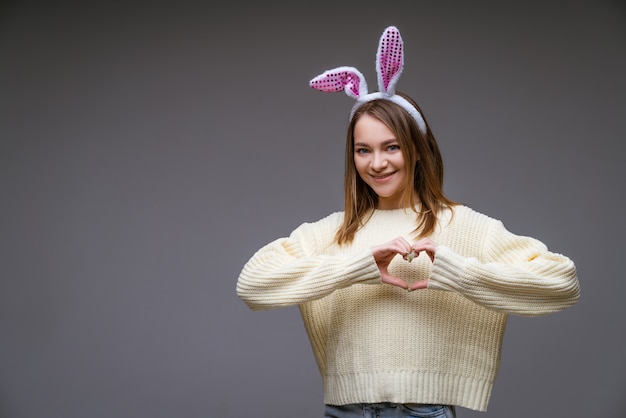 smiling young caucasian girl, blonde with bunny ears, shows a heart with two hands and looking at camera isolated over gray scene