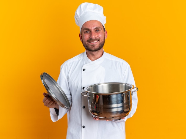 Smiling young caucasian male cook in chef uniform and cap holding pot and pot lid looking at camera isolated on orange wall