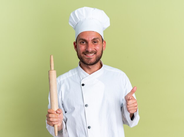 Sorridente giovane maschio caucasico cuoco in uniforme da chef e berretto che tiene mattarello guardando la telecamera che mostra il pollice in alto isolato sul muro verde oliva