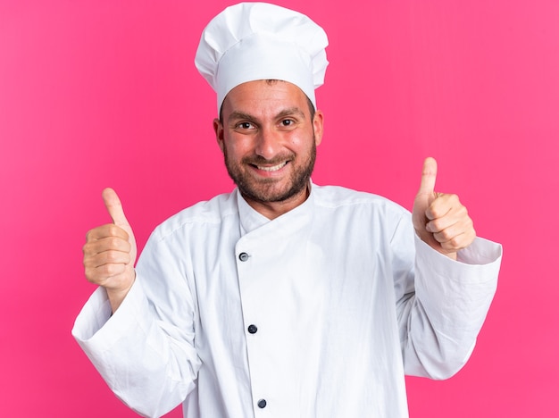 Sorridente giovane maschio caucasico cuoco in uniforme da chef e berretto che mostra i pollici in su