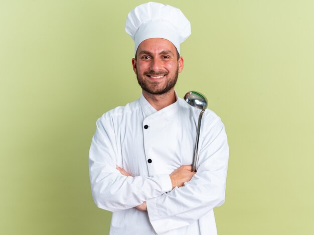 Sorridente giovane maschio caucasico cuoco in uniforme da chef e berretto in piedi con postura chiusa che tiene mestolo guardando la telecamera isolata sul muro verde oliva