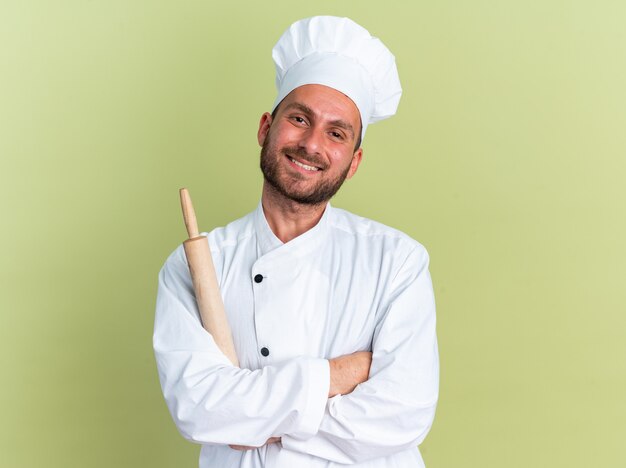 Sorridente giovane maschio caucasico cuoco in uniforme da chef e berretto in piedi con postura chiusa che tiene mattarello guardando la telecamera isolata sulla parete verde oliva con spazio di copia