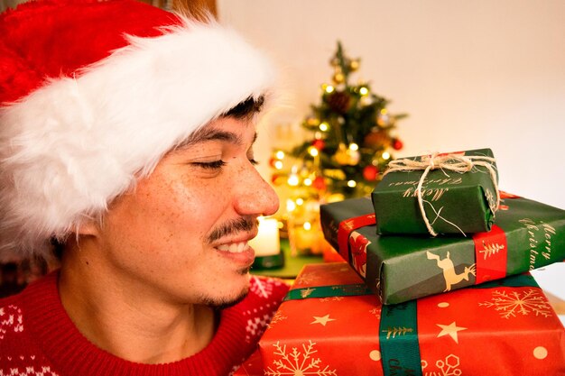 Photo smiling young caucasian man in christmas hat and sweater looking to his side at the christmas gifts