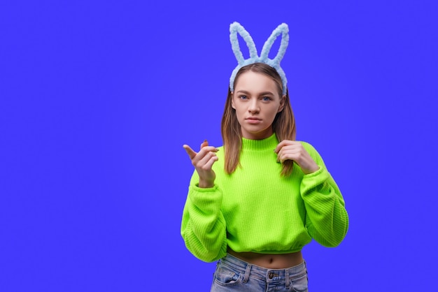 Smiling young caucasian woman, blonde with blue bunny ears and a green sweater, looking at camera isolated over blue wall