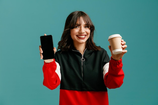 Smiling young caucasian woman showing mobile phone and takeaway coffee cup looking at camera isolated on blue background