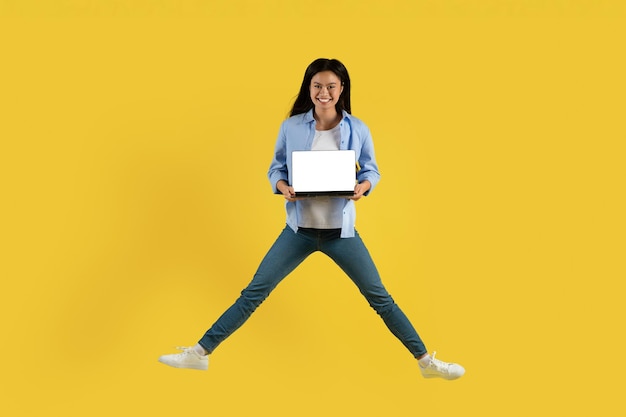 Giovane studentessa cinese sorridente nel salto casuale che mostra il computer portatile con lo schermo in bianco