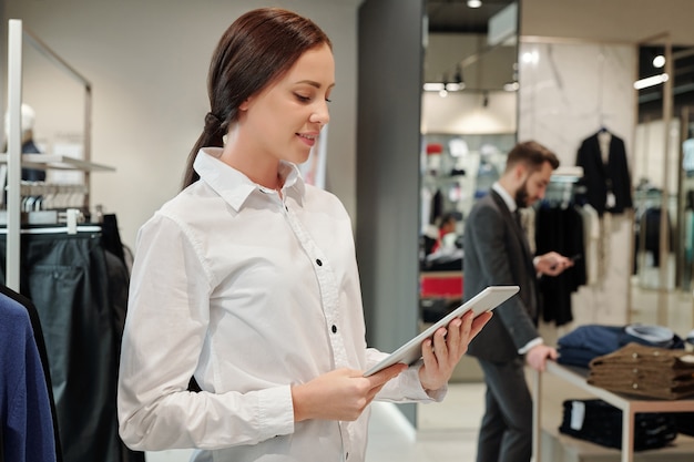 Улыбающийся молодой сотрудник магазина одежды с помощью планшета для поиска кода продавца, пока бизнесмен выбирает одежду