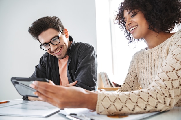 Smiling young colleagues using smartphone together while sitting by the table at office