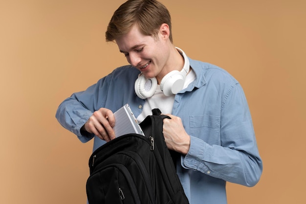 Улыбающийся молодой студент колледжа кладет книги в рюкзак на бежевом фоне