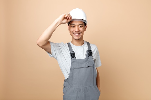 Smiling young construction worker wearing safety helmet and uniform grabbing helmet 