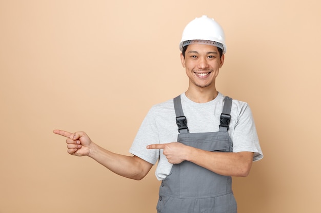 Smiling young construction worker wearing safety helmet and uniform pointing to side 