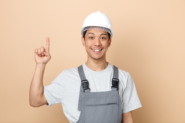 Smiling young construction worker wearing safety helmet and uniform pointing up 