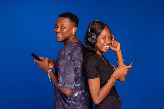 Smiling young couple friends man woman in casual clothes standing back to back using mobile cell phones