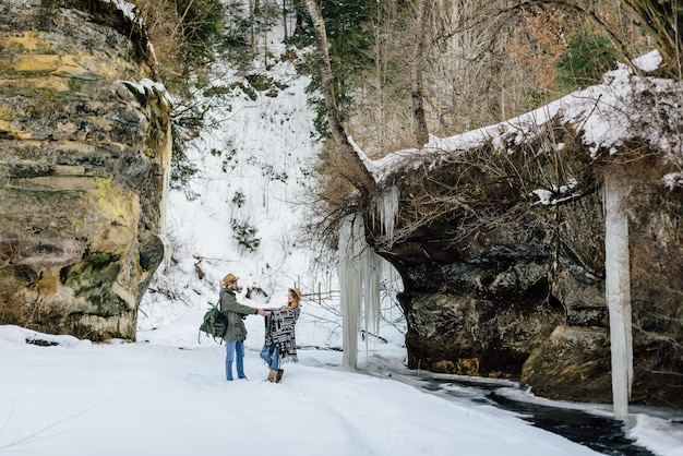 Улыбающаяся молодая пара в туристическом снаряжении, стоящая у частично замерзшей реки, протекающей через заснеженный лес, во время зимнего похода