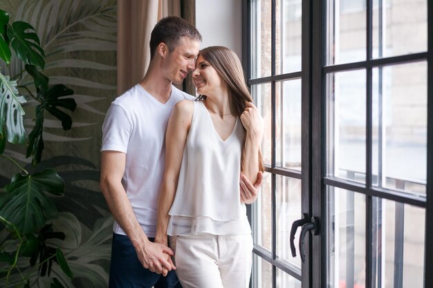 Smiling young couple hugging each other and standing near window looking