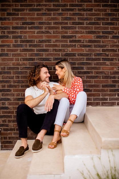 Smiling young couple in love sitting in front of house brick wall