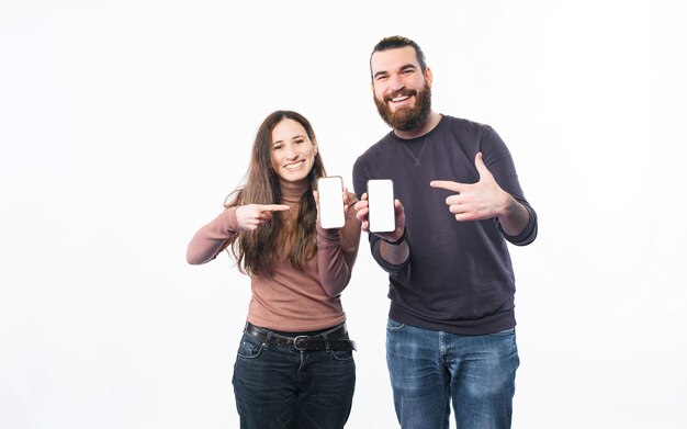 Smiling young couple standing over white wall and pointing at smartphone screens