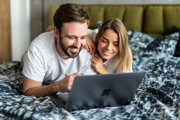 Giovani coppie sorridenti che per mezzo di un computer portatile che si trova sul loro letto