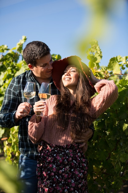 ブドウ園に立っているワインのグラスと笑顔の若いカップル。ブドウ園のスタイリッシュなカップル。