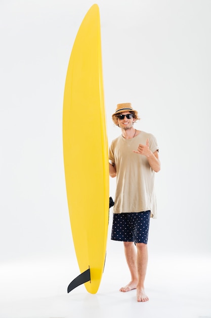 Smiling young curly surfer showing shaka or hang loose sign gesture and holding surfboard isolated on the white wall