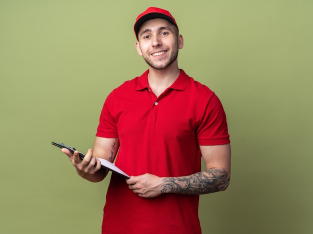 Smiling young delivery guy wearing uniform with cap holding clipboard 