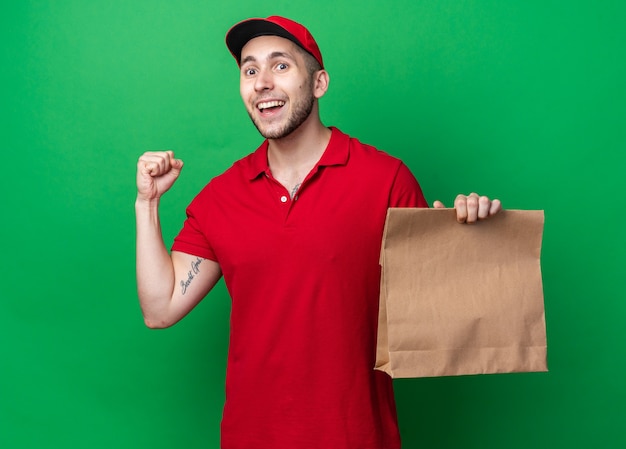 Smiling young delivery guy wearing uniform with cap holding paper food bag showing yes gesture 