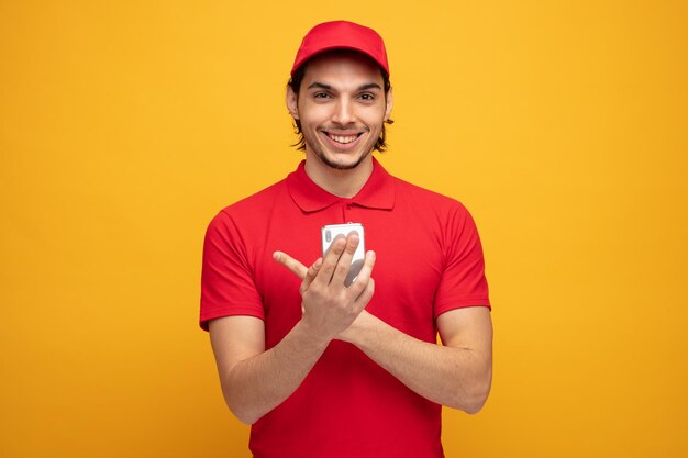 smiling young delivery man wearing uniform and cap holding mobile phone looking at camera pointing to side isolated on yellow background