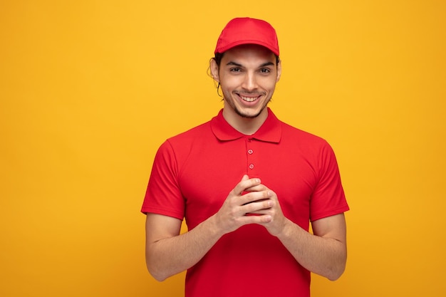 smiling young delivery man wearing uniform and cap keeping hands together looking at camera isolated on yellow background