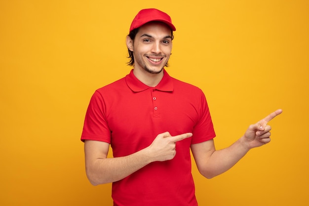 Sorridente giovane fattorino che indossa uniforme e cappuccio guardando la fotocamera che punta al lato isolato su sfondo giallo