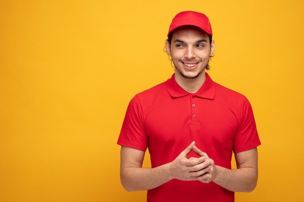 smiling young delivery man wearing uniform and cap looking at side while keeping hands together isolated on yellow background with copy space