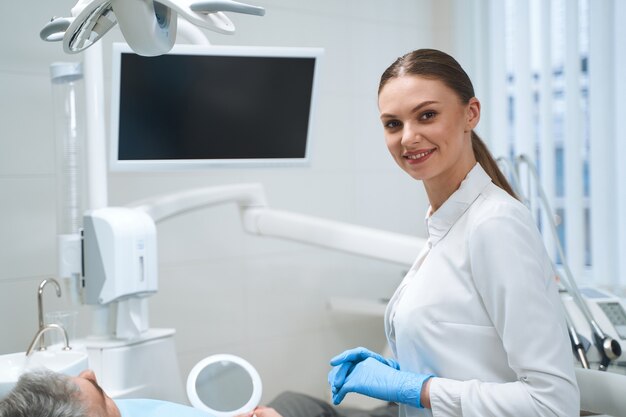 Il giovane medico sorridente in uniforme bianca è in piedi in ufficio accanto allo schermo digitale mentre cura il maschio