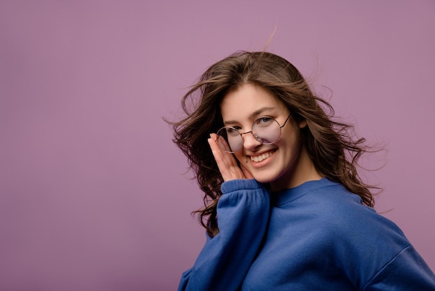 Smiling young european brunette girl in trendy glasses and hoodie looks at camera on purple background People sincere emotions lifestyle concept