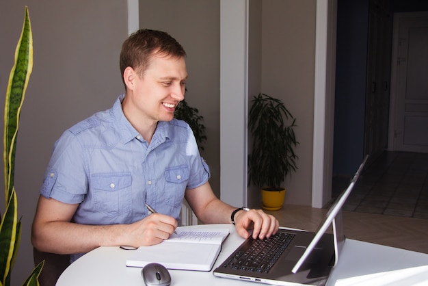 Smiling young European businessman works at home with a laptop and writes with a pen in a notebook