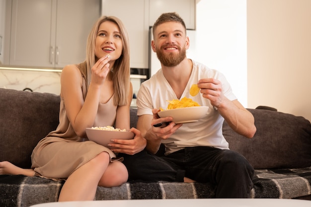 Sorridente giovane coppia europea seduta sul divano e guardare la tv o un film. ragazzo e ragazza che mangiano patatine e popcorn. tempo libero e riposo a casa. concetto di godersi il tempo insieme. interno del monolocale