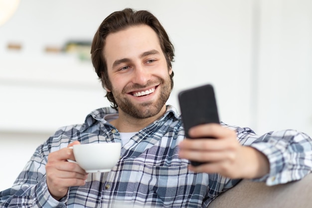 Smiling young european guy with stubble in shirt drinking favorite hot drink and chatting online in