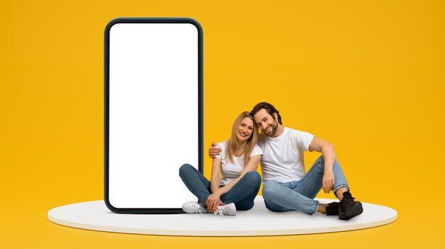 Smiling young european man and female hugging sit on floor with huge smartphone with empty screen