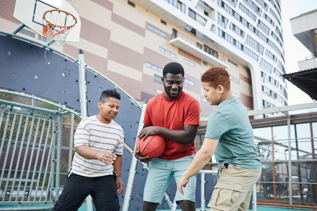 Smiling young father playing basketball with sons and hiding ball from them on city sports ground