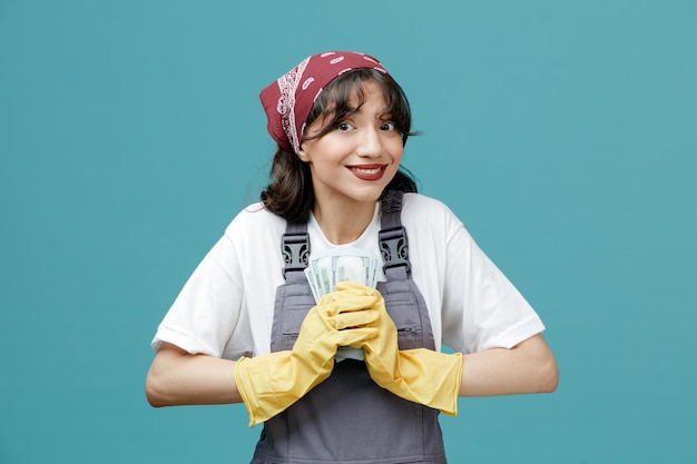 Sorridente giovane donna addetta alle pulizie che indossa bandana uniforme e guanti di gomma che tengono denaro con entrambe le mani guardando la fotocamera isolata su sfondo blu