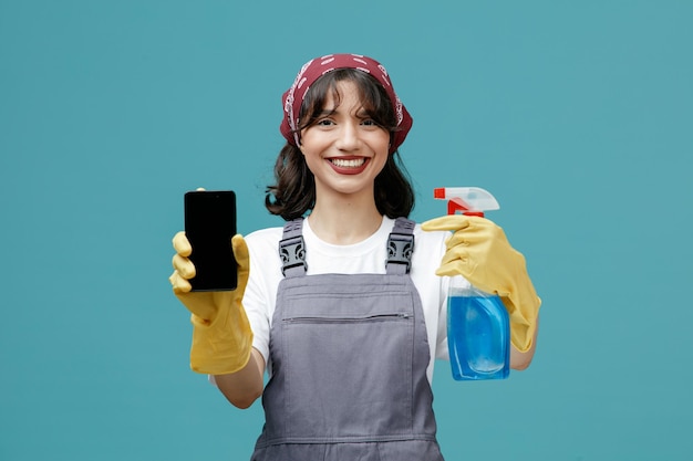 Smiling young female cleaner wearing uniform bandana and rubber gloves showing mobile phone and cleanser looking at camera isolated on blue background