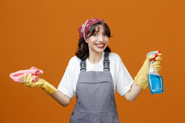 Smiling young female cleaner wearing uniform rubber gloves and bandana holding cleanser and cloth duster looking at camera isolated on orange background