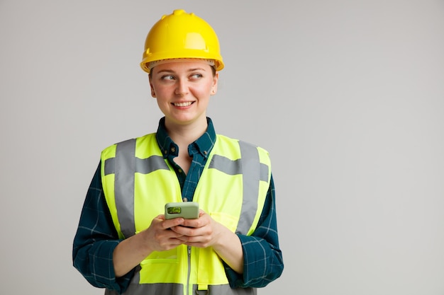 Foto sorridente giovane operaio edile femminile che indossa il casco di sicurezza e giubbotto di sicurezza tenendo il telefono cellulare con entrambe le mani guardando a lato