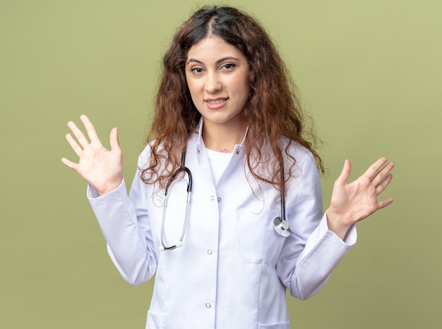 Улыбающаяся молодая женщина-врач в медицинском халате и стетоскопе, смотрящая на фронт, показывает пустые руки, изолированные на оливково-зеленой стене