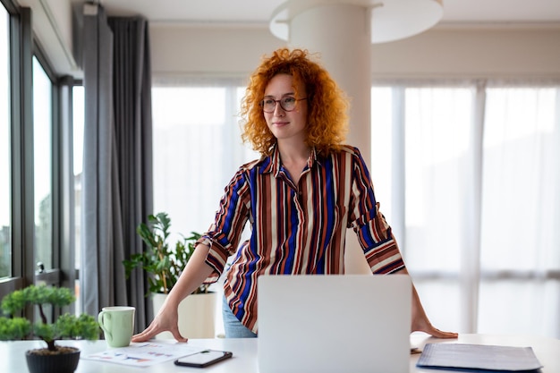 Smiling young female employee stand at desk in office look in\
distance thinking or visualizing career success happy businesswoman\
plan or dream at workplace business vision concept