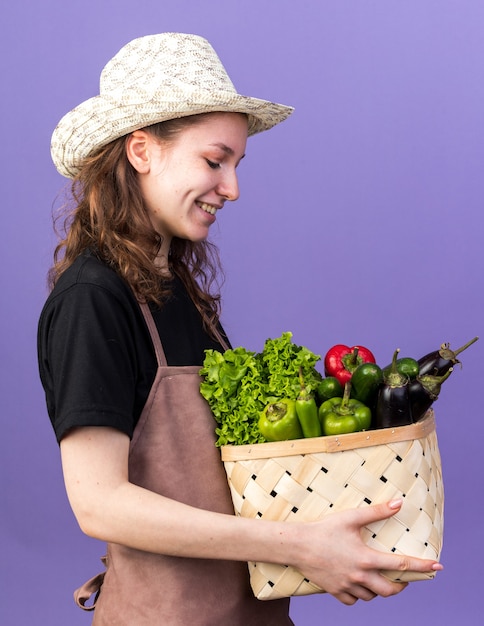 Smiling young female gardener wearing gardening hat holding and looking at vegetable basket isolated on blue wall