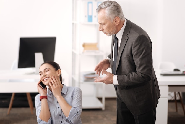 Smiling young female holding telephone near right ear touching her cheek keeping eyes closed