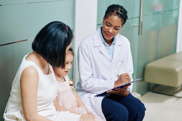 Улыбающаяся молодая женщина-педиатр разговаривает с маленькой девочкой и ее матерью и делает записи в медицинской карте