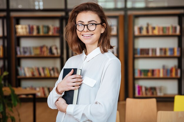 Улыбающаяся молодая студентка в очках держит книгу