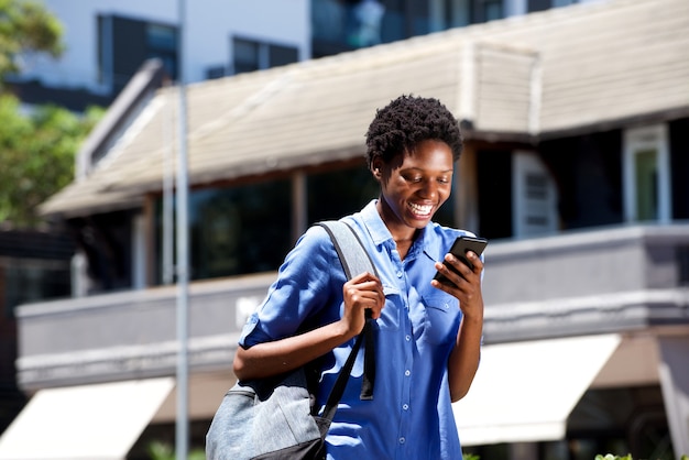 Giovane studentessa sorridente che cammina all'aperto e che legge messaggio di testo sullo smart phone