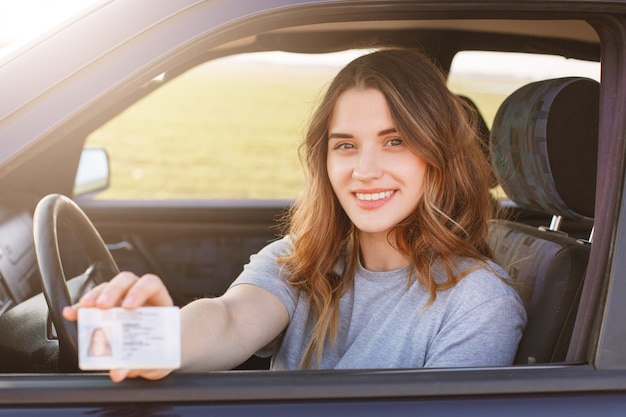 快適な外見で笑顔の若い女性は、運転免許証を誇らしげに示し、新車に座って、未経験の若い運転手であることを示しています