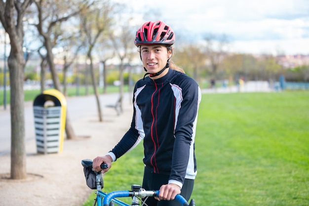 Foto ciclista maschio giovane e sorridente in forma in abbigliamento sportivo e casco seduto sulla bicicletta nel parco